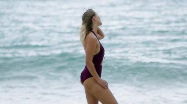 Şık bikinili bir kadın okyanusun kenarında zarif bir şekilde duruyor, sakin ve sakin sahil atmosferinin tadını çıkarıyor.