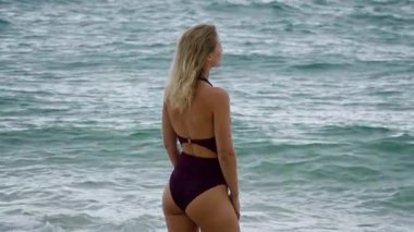 Şık bikinili bir kadın okyanusun kenarında zarif bir şekilde duruyor, sakin ve sakin sahil atmosferinin tadını çıkarıyor.