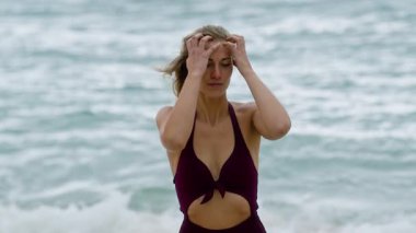 Uzun dalgalı saçları olan kendine güvenen bir kadın gururla okyanus kenarında poz verir. Mor, şık mayosunu sergiler.