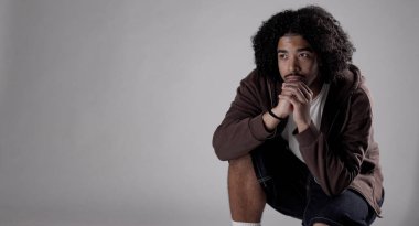 Kıvırcık saçlı, kahverengi kapüşonlu, gri arka planda poz veren genç bir adamın stüdyo portresi. Bu görüntü moda güzelliği ve yaşam tarzı projeleri için uygundur..