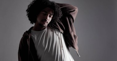 Gri bir arka planda kahverengi kapüşonlu ve beyaz tişörtlü afro saçlı şık bir gencin stüdyo portresi. Bu görüntü moda güzelliği ve yaşam tarzı kavramları için kullanılabilir..