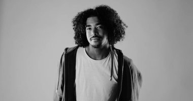 Siyah beyaz kıvırcık saçlı, izole edilmiş bir arka planda tişört ve kapüşonlu genç bir adamın stüdyo portresi. Bu görüntü moda ve güzellikle ilgili projeler için uygundur..