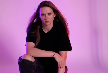 Uzun saçlı, siyah tişörtlü ve pembe arka planda yırtık kot pantolon giyen güzel bir kadının stüdyo portresi. Bu görüntü moda ve güzellik projeleri için uygundur..