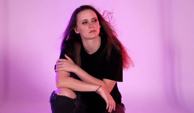 Uzun saçlı, siyah tişört giyen ve pembe arka plana poz veren yırtık kotlu genç bir kadının stüdyo portresi. Bu görüntü moda güzelliği ve yaşam tarzı projeleri için uygundur..