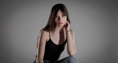 Uzun saçlı, siyah kolsuz bluz ve gri arka planda kot giyen esmer bir kadının stüdyo portresi. Bu görüntü moda ve güzellik projeleri için uygundur..