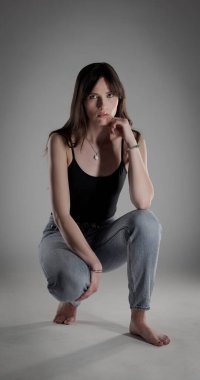 Siyah kolsuz bluz giyen esmer bir kadının stüdyo portresi ve gri arka planda çömelmiş kot pantolon. Bu görüntü moda ve güzellik projeleri için uygundur..
