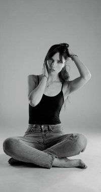 Tek renkli stüdyo portresi, kot pantolon giymiş ve kolsuz tişörtlü gri bir arka planda oturan genç bir kadın. Bu moda ve güzellik malzemesi fotoğrafçılığı çeşitli ticari kullanımlar için mükemmeldir..