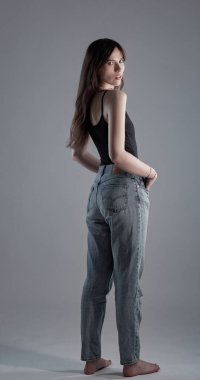 Kot pantolonlu ve siyah kolsuz tişörtlü bir kadının gri bir arka planda omzunun üzerinden baktığı bir fotoğraf. Bu görüntü moda güzelliği ve yaşam tarzı projeleri için uygundur..