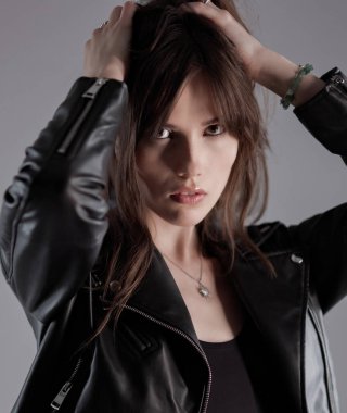 Modaya uygun esmer bir mankenin stüdyo portresi. Siyah deri ceket giyiyor. Gri arka planda elleri saçında. Bu görüntü güzellik modası ve tarz kavramları için kullanılabilir..