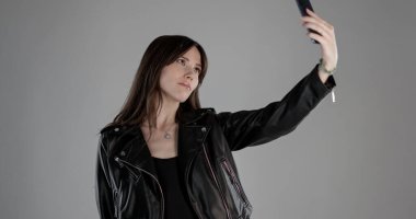 Siyah deri ceketli esmer bir kadının gri arka planda akıllı telefonuyla selfie çektiği bir fotoğraf. Bu görüntü moda güzelliği ve yaşam tarzı içeriği için uygundur..