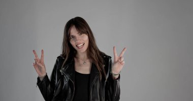 Siyah deri ceketli, gülümseyen esmer bir kadının gri arka planda barış işareti yaptığı bir fotoğraf. Bu görüntü moda ve güzellik reklam kampanyaları için uygundur..