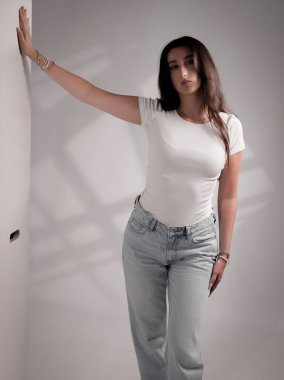 Beyaz tişört ve kot pantolon giymiş beyaz bir duvara poz veren genç bir kadının stüdyo fotoğrafı. Bu görüntü moda güzelliği ve yaşam tarzı reklam kampanyaları için uygundur..
