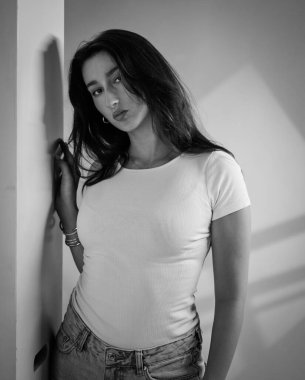 Tek renkli stüdyo resmi beyaz tişört ve kot pantolon giymiş duvara dayanmış genç bir kadın. Bu görüntü moda güzelliği ve yaşam tarzı projeleri için uygundur..