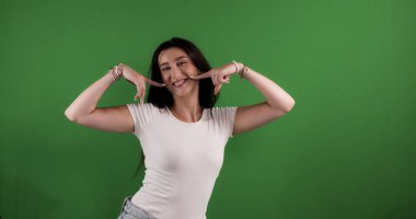 Stüdyoda gülümseyen esmer bir kadının yeşil arka planda poz verirken yanaklarında parmakları olan bir fotoğrafı. Bu resim güzellik ve ürün modelleme kavramları için uygundur..