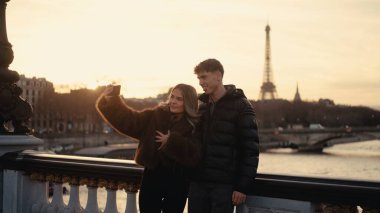 Genç bir çift gün batımında Paris 'te arka planda Eyfel Kulesi ile selfie çekiyor. Bu resim seyahat turizmi ve romantizmle ilgili içerik için uygundur.