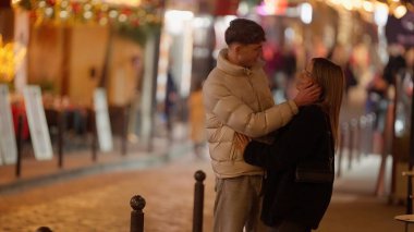 Romantik bir çift gece Paris 'te bir sokakta sarılır ve etrafı bokeh ışıklarıyla çevrilidir. Bu görüntü, sevgililer günü veya seyahat içeriği için uygun bir şehir ortamında sevgi ve şefkat yakalar..