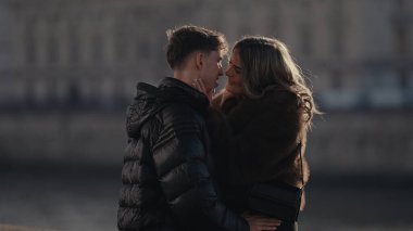 Paris 'teki romantik bir çift, güneşli bir günde Seine Nehri yakınlarında kışlık palto ve ceket giyerken sevgi ve şefkat dolu bir anı paylaşıyor. Bu resim seyahat turizmi veya ilişki temalı kampanyalarda kullanılabilir.