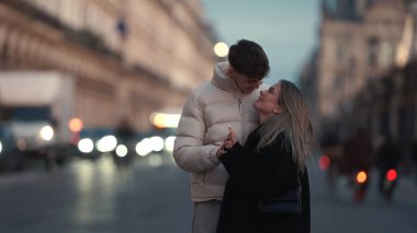 Birbirine aşık genç bir çift, alacakaranlıkta Paris sokaklarında el ele tutuşurken, arka planda şehir ışıkları ve binalar bulanıklaşıyor. Bu resim, romantizm seyahatleri veya kentsel yaşam tarzı kavramlarını resmetmek için ticari olarak kullanılabilir..
