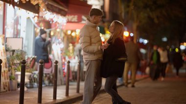 Romantik bir çift gece Paris sokaklarında el ele tutuşuyor. Etrafı bokeh ışıkları ve şenlikli süslemelerle çevrili. Bu resim Sevgililer Günü seyahat kavramları ya da şehir ortamında aşk ve ilişkileri resmetmek için uygundur..