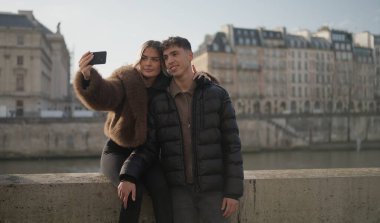 Romantik bir çift Paris 'te akıllı telefonuyla selfie çekiyor. Fransa' da Seine Nehri 'nde ve arka planda güneşli bir günde binalar var. Bu resim seyahat ve turizmle ilgili içerik için uygundur.