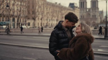 Genç bir çift, Paris 'te bir şehir caddesinde sevgiyle kucaklaşır. Arka planda Notre Dame Katedrali görünür. Görüntü, seyahat romantizmi ya da yaşam tarzı memnuniyeti için uygun bir aşk ve bağlantı anını yakalar..