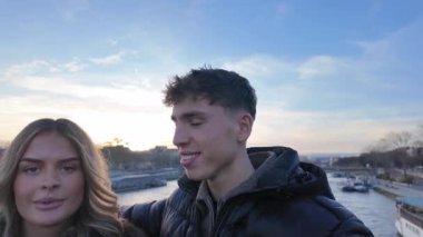 Paris 'te bir çift Seine Nehri' nde selfie çekiyor ve arka plandaki şehir manzarası elde taşınabilir POV tarzında çekiliyor. Bu da ona samimi ve doğal bir sosyal medya içeriği veya seyahat vlog 'u veriyor..