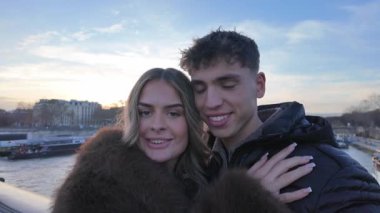 Paris 'te bir çift gün batımında Seine Nehri' nin üzerindeki bir köprüde samimi selfie çekiyorlar. Video, çiftin poz verirken elde çekilmiş bir görüntüsünü ve Avrupa 'da romantik bir tatil sırasında sosyal medya içeriği oluşturan kameraya gülümsemelerini gösteriyor..