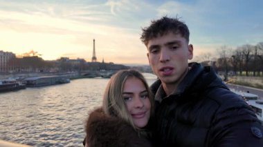 Paris 'te bir çift arka planda Eyfel Kulesi ile samimi selfie çekiyor. Video günbatımında Seine Nehri ile kucaklaşan ve gülümseyen çiftin el yapımı POV fotoğrafını gösteriyor..
