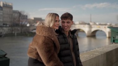 Romantik bir çift Paris 'te Seine Nehri kıyısında sarılır kadın kürk giyer ve erkek kabarık bir ceket giyer güneşli bir günde bir köprünün yanında durup Avrupa seyahatlerinin ve tatillerinin tadını çıkarırlar..