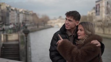 Bir çift Paris 'teki Seine Nehri' nin üzerinde dururken sarılıyor ve işaret ediyor. Bu sahne, Avrupa 'da seyahat aşklarını ve turizmi vurguluyor..