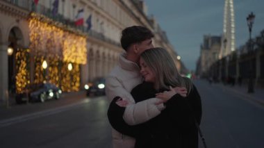 Romantik bir çift, akşam karanlığında Paris 'te Fransa' nın bir caddesinde sarılırken görülüyor. Eyfel Kulesi arka planda görünüyor ve sahne yumuşak, sıcak ışıklarla aydınlatılıyor..