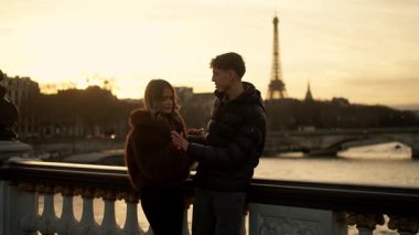Romantik bir çift Paris 'te bir köprüde duruyor. Eiffel Kulesi arka planda gün batımında görünüyor. Altın saat ışığı ortamı aydınlatıyor ve sıcak ve sevgi dolu bir ortam yaratıyor.