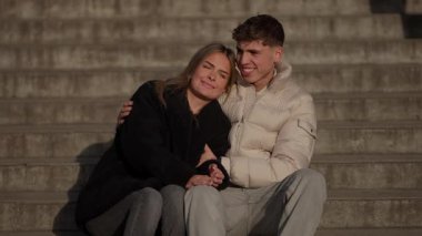 Genç bir çift Paris 'te beton basamaklara oturur kadın erkeğin omzuna yaslanır altın saat ışığı sıcak romantik bir atmosfer yaratır aşk ve beraberliği Avrupa kentsel ortamında yakalar.