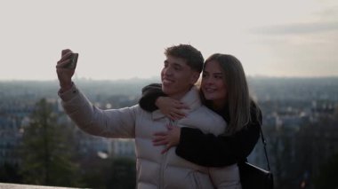 Genç bir çift Paris 'te arka planda şehir manzarası olan bir selfie çeker. Erkek akıllı telefonu tutarken kadın onu kucaklar ve birlikte geçirdikleri zamanın tadını çıkarır..