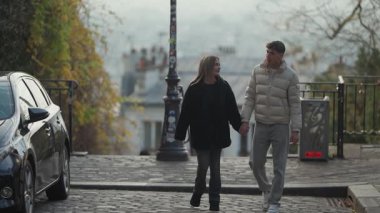Genç bir çift, Paris 'te bir kaldırım taşı caddesinde el ele yürür. El kamerası, arka planda manzaralı şehir manzarasıyla romantik yürüyüşlerini yakalar..