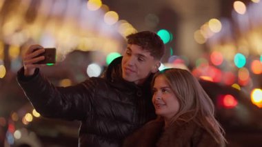 Genç bir çift gece Paris 'te Fransa' da selfie çekiyor. Erkek ve kadın gülümsüyor ve akıllı telefondan poz veriyorlar. Bokeh ışıkları şehirde şenlikli ve romantik bir atmosfer yaratıyor..