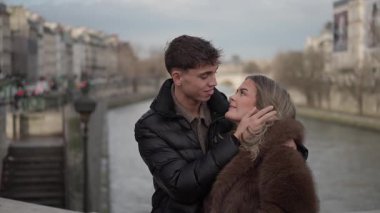 Romantik bir çift Paris 'te, Fransa' da Seine nehri ve arka plandaki binalarla kucaklaşır genç adam ve kadın bir köprüde kışlık giysiler giyerken sevgi ve sevgi gösterirler..