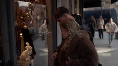 Paris 'te bir çift mağaza vitrinine bakıyor. Pencerede 30 indirim tabelası ve beyaz tavşan süsü var. Arka planda yürüyen insanlar var..