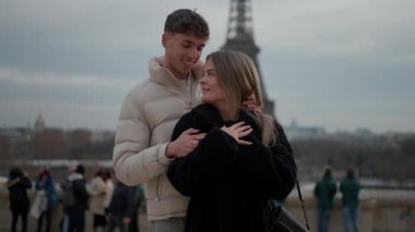 Genç bir çift Paris 'te arka planda Eyfel Kulesi ile kucaklaşıyor. Erkek ve kadın bulutlu bir günde birbirlerine sevgi ve sevgiyle bakıyor. Bu sahne ikonik şehirde romantik bir anı yakalıyor..