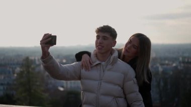 Genç bir çift arka planda Paris silueti olan bir selfie çekiyor erkek ve kadın Fransa 'daki seyahat anılarını yakalamak için bir akıllı telefon kullanıyorlar..