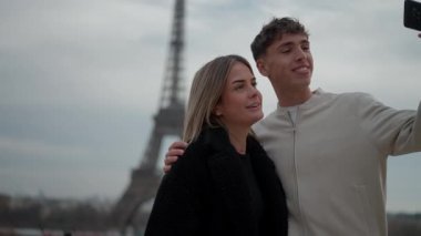 Genç bir çift, Paris 'teki Eyfel Kulesi' nde bir selfie çekiyor. Fransa 'da erkek ve kadın, arka planda ikonik simge olan bir fotoğraf için birbirlerine sarılıp gülümsüyorlar.
