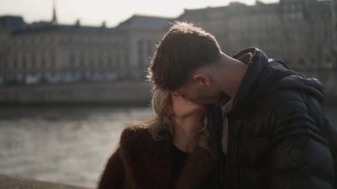 Paris 'te Seine nehri ve şehir binaları arka planda romantik bir çift öpüşür. Sahne, sığ bir alan derinliğiyle Avrupa kentsel ortamında sevgi ve şefkat yakalar..