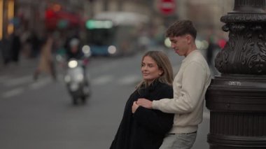 Genç bir çift, Paris 'te bir sokak lambasının yanında sarılıyor. Arka planda şehir trafiği bulanıklaşıyor. Bu sahne, bir Avrupa kentsel ortamında sevgi ve şefkat anı yakalıyor..