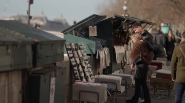 Romantik bir çift Paris 'teki Seine Nehri' nin kıyısındaki kitapçıda kitaplara göz gezdirirken görülüyor. Sahne, arka planda sevgili kitapçıları ve şehrin ikonik mimarisi ile Paris yaşamının özünü yakalıyor..