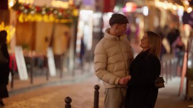 Genç bir çift gece büyüleyici bir Paris caddesinde kucaklaşır. Sıcak sokak lambaları ve şenlikli dekorasyonlar altında birbirlerine sevgiyle bakarlar. Sığ alan derinliği romantik bir bokeh etkisi yaratır..