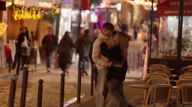 Genç bir çift, Paris 'in bir sokağında, yumuşak bir bokeh ve sıcak bir aydınlatmayla romantik bir atmosfer yaratıyor. Arka planda yürüyen insanlar ve restoran masaları yakınlarda görünüyor..