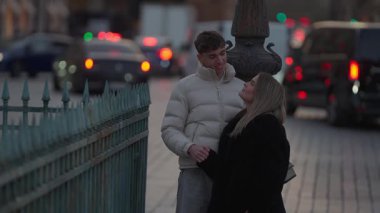 Genç bir çift Paris 'te alacakaranlıkta Fransa' nın bir caddesinde kucaklaşıyor. Arka planda arabalar geçiyor ve sahne sokak lambalarıyla aydınlatılıyor..