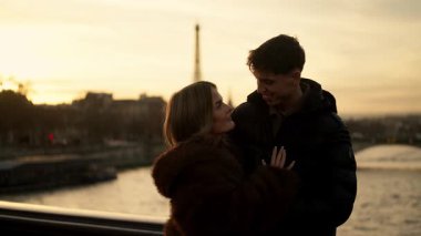 Genç bir çift, Paris 'te bir köprüde Eiffel Kulesi' nin arka planda görülebildiği altın bir günbatımında birbirlerine sarılıyorlar..
