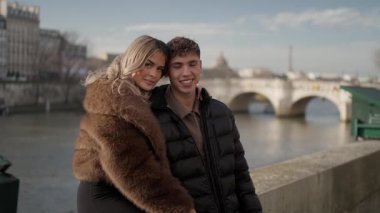 Genç bir çift Paris 'te bir portre için poz veriyor Seine nehri ve köprüleri arka planda görünüyor kadın ve erkek gülümsüyor ve kentte geçirdikleri zamanın tadını çıkarıyorlar..