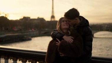 Genç bir çift Paris 'te bir köprüde Eiffel Kulesi' ni arka planda gün batımında görülebilen bir şekilde kucaklaşır. Seine Nehri aşıklar romantik bir anı paylaşırken ve şehrin tadını çıkarırken altından akar..